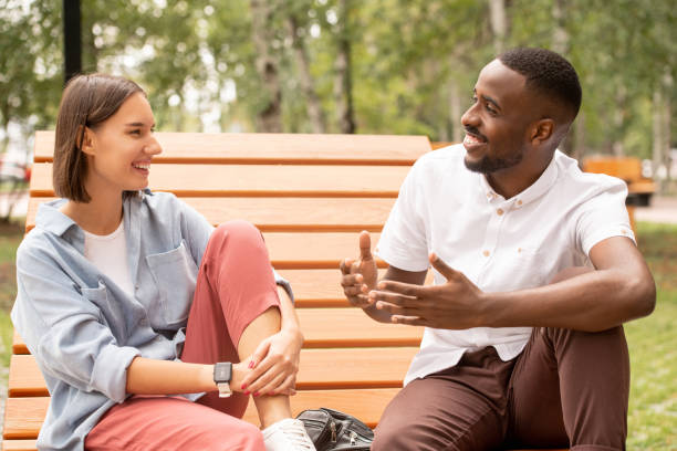 Happy young intercultural couple in casualwear sitting on wooden bench in park or square, chatting and enjoying summer day 
